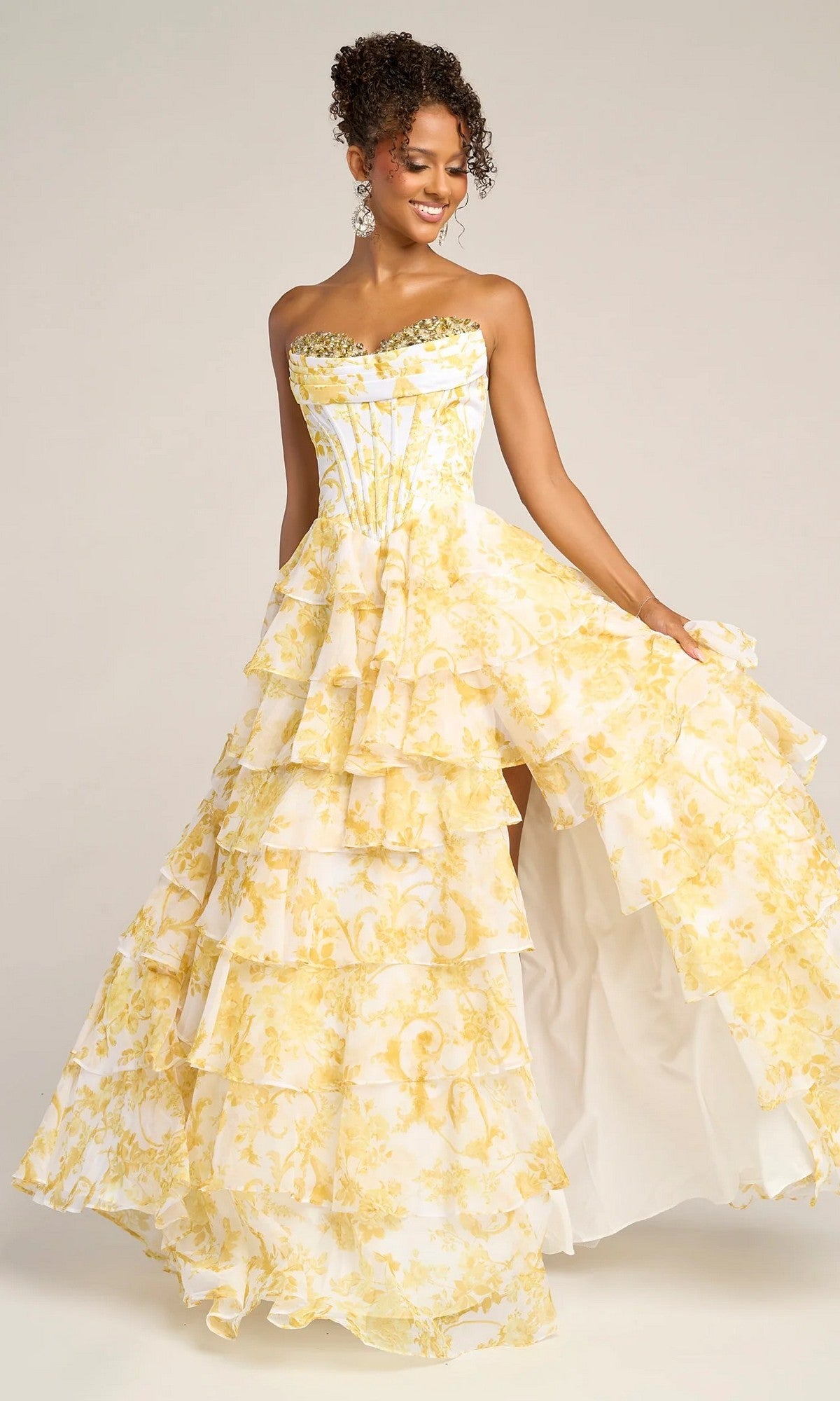 A woman stands smiling, holding out the side of her Portia and Scarlett PS26210 long prom dress—this strapless gown features yellow and white ruffled tiers with floral patterns.